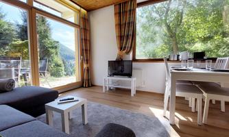 A bright living room with large windows and a view of nature. There is a sofa, a table, and a TV in a modern, inviting space.