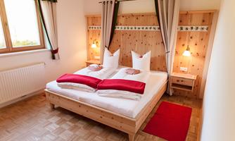 A cozy bedroom with a large double bed and wooden decor. There are bright curtains and a red carpet on the floor.