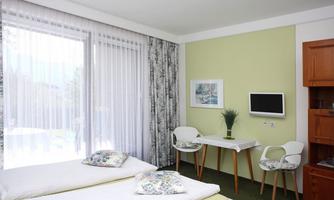A bright room with two single beds and a small table. Large windows let in a lot of light and offer a view of the surroundings.