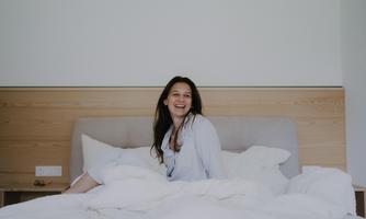 A smiling woman is sitting in bed, enjoying the moment. The surroundings are bright and friendly, with white bedding and a minimalist design.
