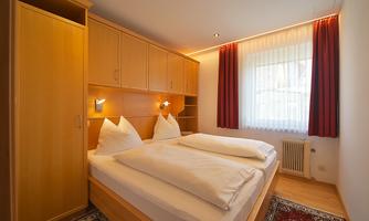 A modern bedroom with two beds and beige wooden furniture. The windows let in plenty of daylight and the walls are painted light.