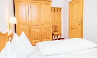 A cozy room with wooden furniture and a bed. The room features a wardrobe, a chair, and a warmly lit atmosphere.