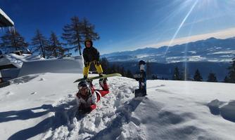 Two children are playing in the snow on a mountain. The surroundings are sunny with a view of the mountains in the distance.