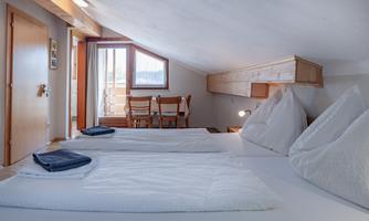 A bright, cozy bedroom with two beds and small swivel chairs. Through the window, the view of a balcony and a dining area is visible.