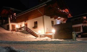 A cozy chalet, surrounded by snow, illuminated at night. The facade showcases traditional wooden details and an inviting atmosphere.