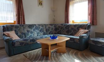 A cozy living room with a blue sofa and a wooden coffee table. The room has large windows and bright curtains.