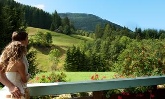 A couple stands on a balcony overlooking a picturesque landscape with green meadows and trees. In the background, gentle hills and a blue sky can be seen.