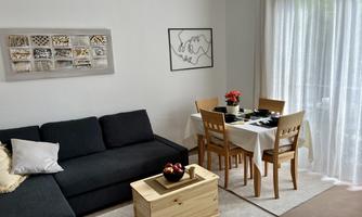A cozy living room with a dark gray sofa, a wooden table, and chairs. Large windows let in plenty of light, and the decoration is modern and inviting.
