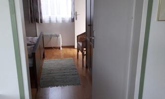 A bright hallway with a wooden floor and an open door to a room. A small rug and a chair are visible in the room.