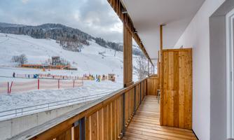 A balcony with a wooden railing and a view of snowy slopes. In the background, skiers and mountain scenery can be seen.