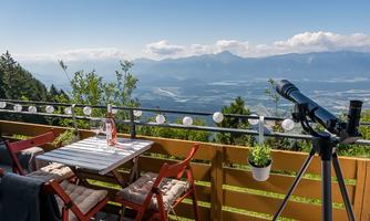 A cozy terrace with red furniture and a view of the mountains. In the background, green hills and a clear sky can be seen.