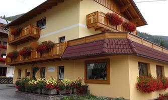 A light yellow building with wooden cladding and balconies, adorned with red flowers. In front of the house, there is a flower garden with colorful plants.