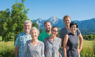 A group of six people stands in front of a picturesque mountain landscape. The sky is clear and the surroundings are green and inviting.