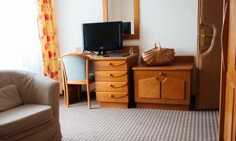 A cozy hotel room with a bed, a table, a TV, and a large window. The furniture is made of wood, and the floor is covered with a checkered carpet.