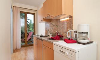 A small, modern kitchen with wooden cabinets and a dining area. The view of a beam with chairs is visible.