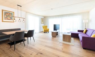 A modern living room with light wooden flooring, a purple couch, and a dining area. Large windows let in plenty of daylight.