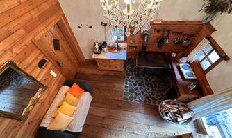 A cozy wooden house with a modern chandelier. The interior features a seating area with colorful cushions and a kitchen with a rustic wooden table.