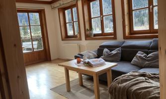 A cozy living room with a gray sofa and a wooden table. Large windows offer a view of nature outside.