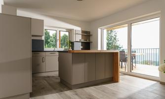 A modern kitchen with light cabinets and a wooden worktop. The large windows offer a view of the outdoor area and plenty of natural light.