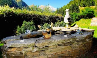 A beautiful garden with a stone grill and a small decoration. In the background, green mountains and a clear blue sky are visible.
