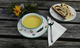 Eine Schale mit heller Suppe steht auf einem Holztisch, umgeben von einem bunten Blumenstrauß. Daneben liegt ein Teller mit einem Stück Marmorkuchen und einem Löffel.