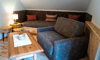A cozy seating area with a brown sofa, a wooden table, and decorative cushions. Next to the sofa is a lamp, and there are small wooden tables.