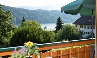 A beautiful view from a balcony over a lake and green trees. In the foreground, there is a table with flowers and a sunshade.