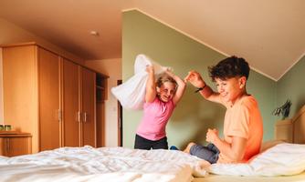Two children are happily playing on a bed and having a pillow fight. The room is bright and cheerfully furnished.