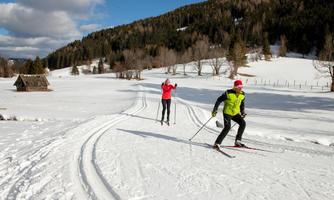 Two cross-country skiers are traveling through a snowy landscape. In the background, forests and hills can be seen.