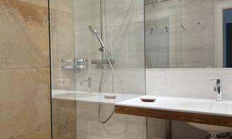 A modern bathroom with a glass shower and a wooden shelf. The walls are light and made of fine tiles.