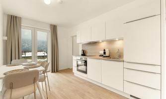 A modern kitchen with white furniture and wooden floors. Large windows let in plenty of light and provide a view outside.