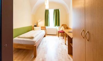 A simply furnished room with a single bed, a desk, and a chair. The windows are covered with green curtains and the floor is made of light wood.