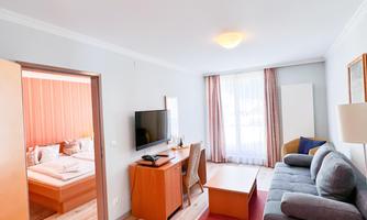 A bright room with a sofa, a table, and a television. In the background, there is a bedroom with a bed.