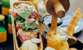 An appealing wooden board with various delicacies, including sausage, cheese, and fresh herbs. There are also small jars of jam and fruit present.