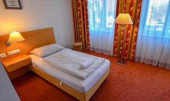 A simple single room with a bed, bedside table, and lamp. The windows are decorated with curtains and the carpet is red.