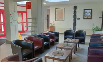 A modern waiting room with comfortable chairs and a table. The walls are bright and light streams through large windows.