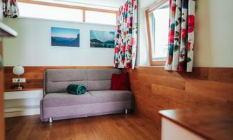 A cozy living space with a gray sofa and a red cushion. Pictures hang on the walls and the windows are decorated with colorful curtains.