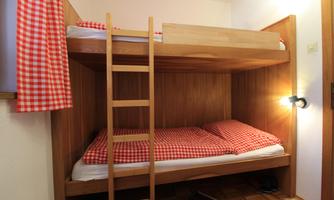 A cozy wooden bunk bed with red checkered bedspreads. The walls are simple and bright, and there is a small ladder to the upper bed.