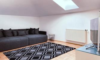 A modern living room with a gray sofa and a rug. There is a television and plenty of daylight coming through the skylight.