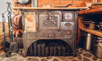 A traditional, rustic metal wood stove in a cozy kitchen. The surroundings are warm and inviting, with various kitchen utensils and pots.