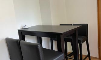 A small dining area with a high table and four black chairs. The wall is light and the floor is made of wood.