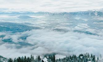 A snowy landscape with houses and snow-covered trees. A cloud cover floats over the mountains.