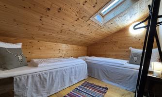 A cozy attic room with two single beds and wooden walls. The decoration includes cushions and a colorful carpet.