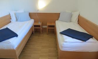 A simple bedroom with two single beds and wooden details. The walls are light, and there is a nightstand with a lamp.