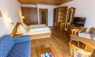 A cozy hotel room with two single beds, a sofa, and a wooden table. The decor is simple and friendly, featuring warm wood and bright lighting.
