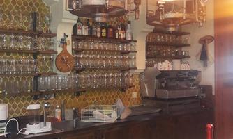 A cozy bar with wooden shelves full of glasses and bottles. In the background, there is a coffee machine and a stylish lamp provides warm light.