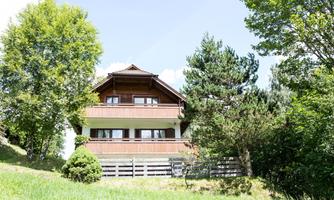 An idyllic wooden house surrounded by trees and meadows. The sky is clear and sunny.