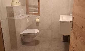 A modern bathroom with gray tiles and light wood details. It features a sink, a toilet, and a window.