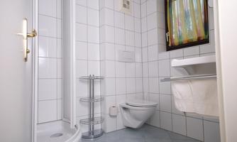 A modern bathroom with white tiles and a shower area. There is a toilet and a shelf for towels.
