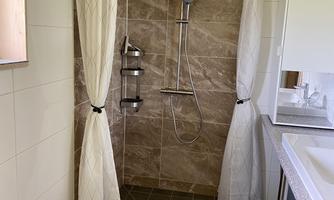 A modern bathroom with a shower and white curtains. The walls are tiled in warm brown tones, and there is a large mirror as well as a bright washbasin.